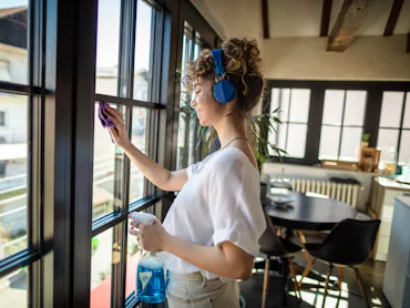 Photo of young female cleaning window in her apartment with headphones.