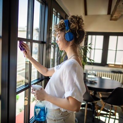 Photo of young female cleaning window in her apartment with headphones.