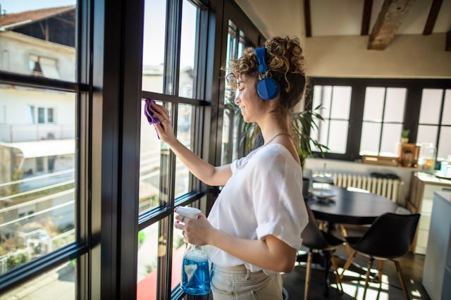 Photo of young female cleaning window in her apartment with headphones.