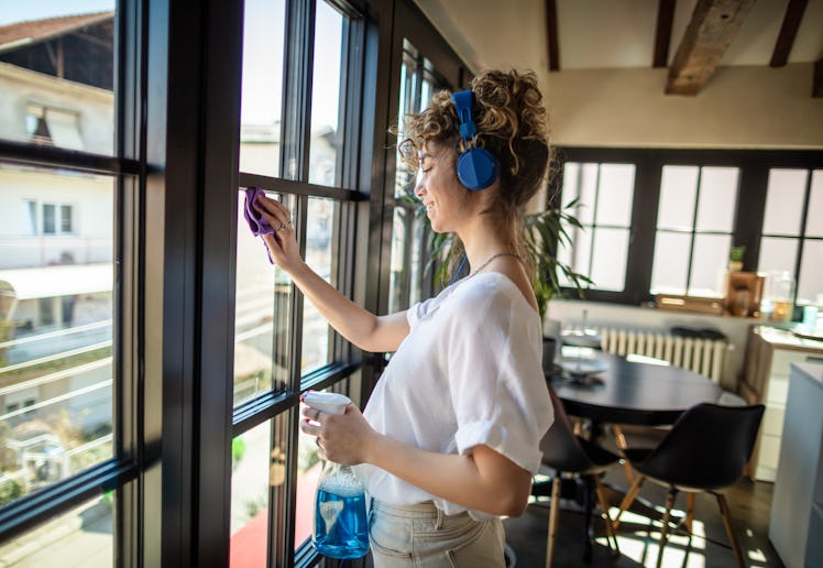 Photo of young female cleaning window in her apartment with headphones.