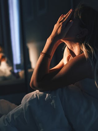 A woman sits on a bed in a dimly lit room, hands covering her face in distress while the mirror show...