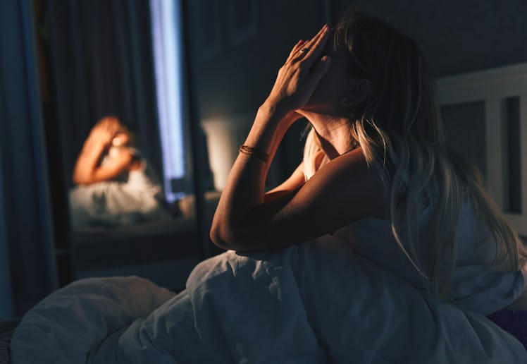 A woman sits on a bed in a dimly lit room, hands covering her face in distress while the mirror show...