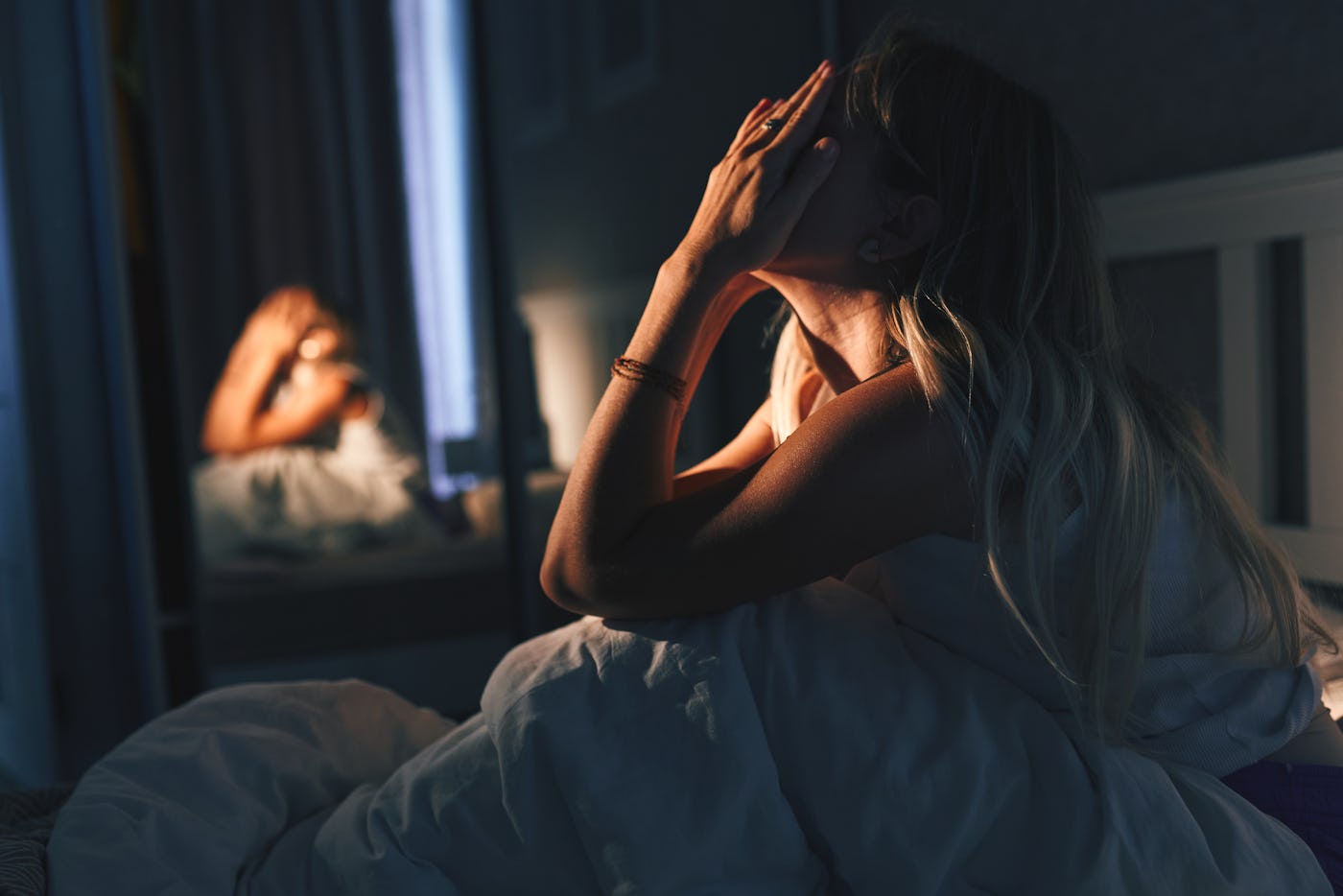 A woman sits on a bed in a dimly lit room, hands covering her face in distress while the mirror shows her blurred reflection. The scene suggests insomnia, worry, and emotional strain.