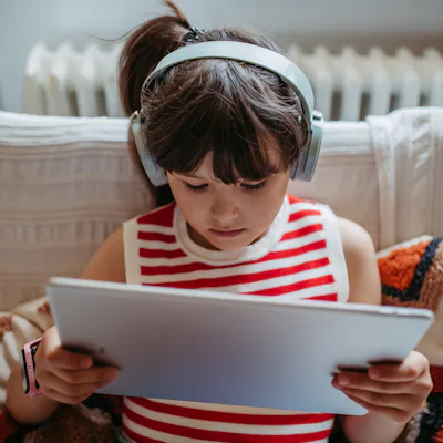 Portrait of a young girl watching a video on a tablet with wireless headphones on. Girl spending her summer indoors, online, and alone. Schoolgirl sitting at home, connected to an online class, embracing remote learning.