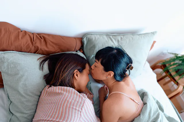 Two adult women, one giving a gentle forehead kiss to the other while lying close in bed under light...