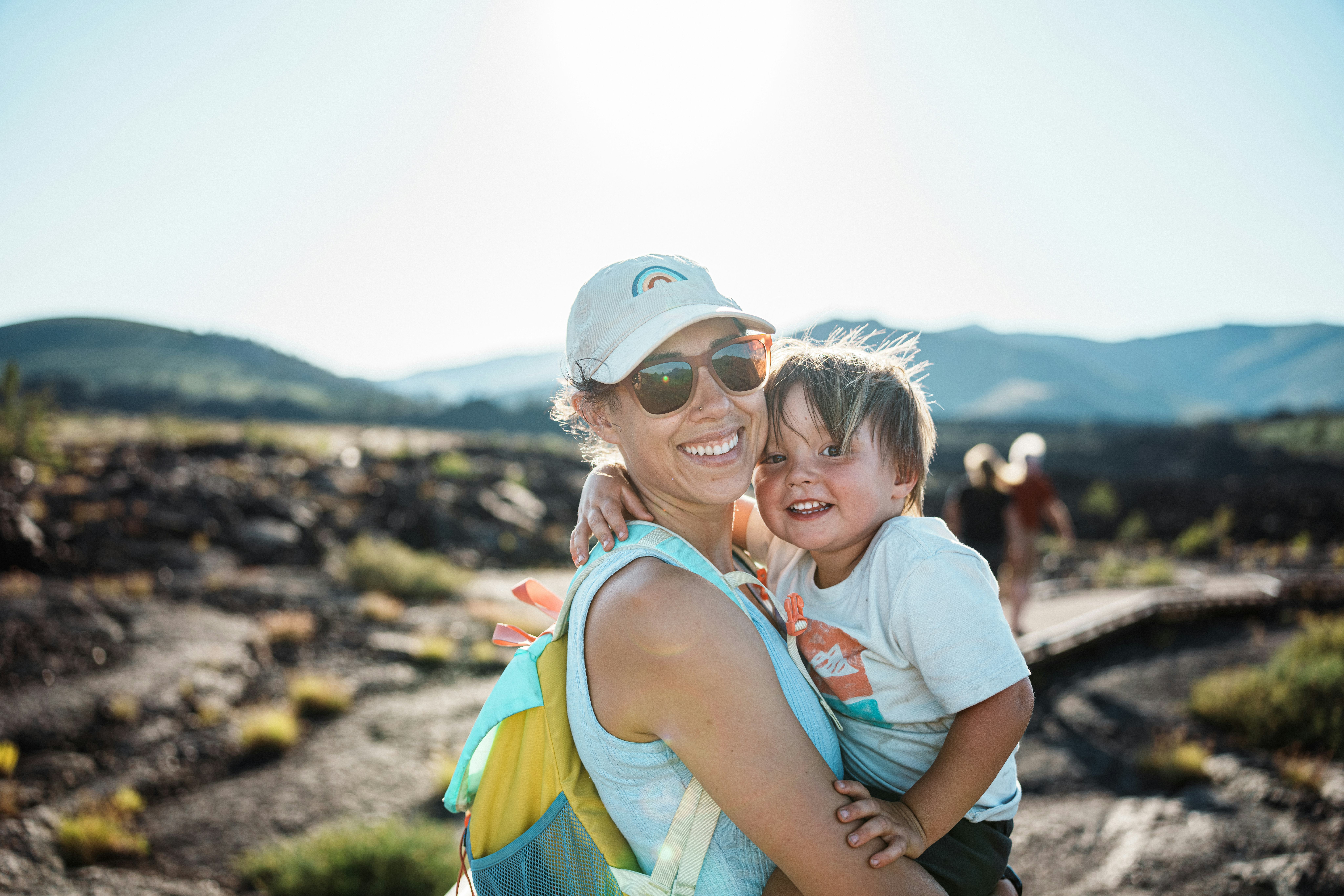 A Eurasian woman affectionately holds her two-year-old son as they smile toward the camera while hik...
