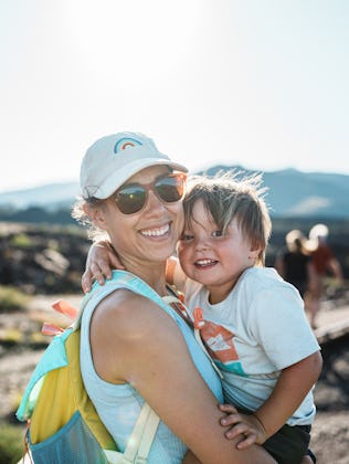 A Eurasian woman affectionately holds her two-year-old son as they smile toward the camera while hik...