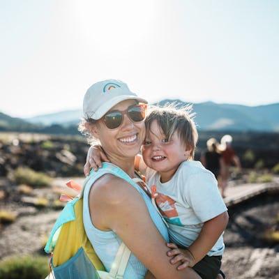 A Eurasian woman affectionately holds her two-year-old son as they smile toward the camera while hiking at Craters of the Moon National Monument in Idaho on a warm and sunny day.