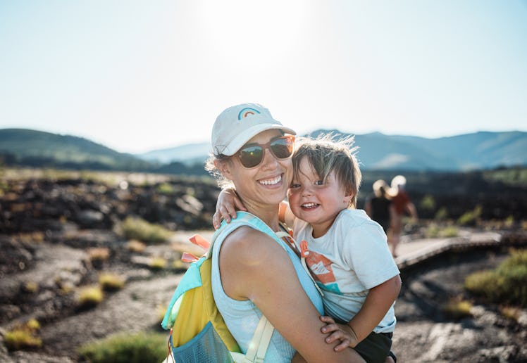 A Eurasian woman affectionately holds her two-year-old son as they smile toward the camera while hik...