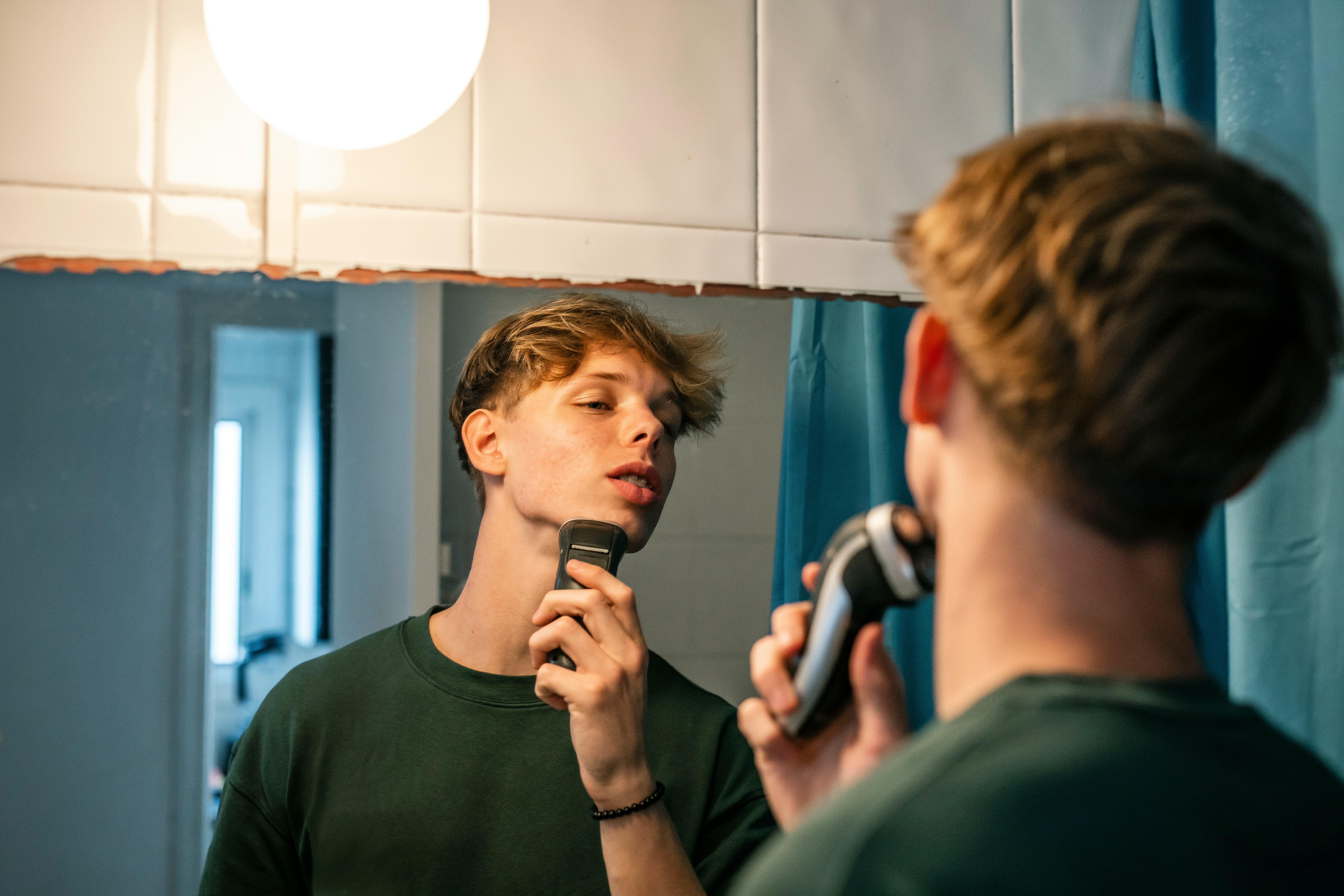 16-year-old young man shaves with a razor in front of the mirror in the bathroom. He is a handsome f...