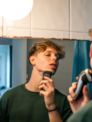 16-year-old young man shaves with a razor in front of the mirror in the bathroom. He is a handsome f...