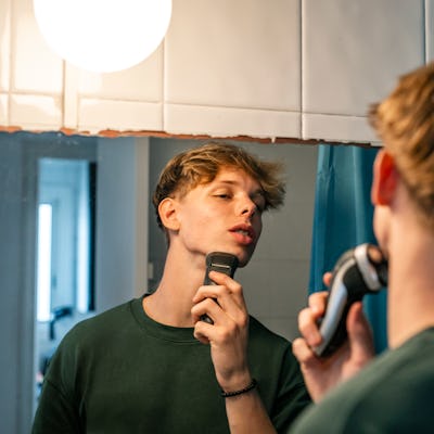 16-year-old young man shaves with a razor in front of the mirror in the bathroom. He is a handsome fair-haired boy.