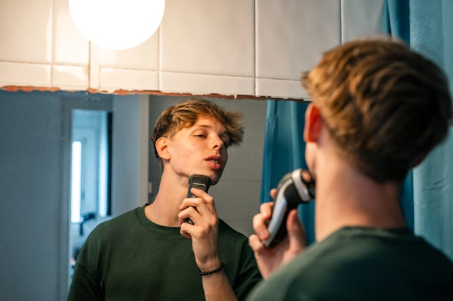 16-year-old young man shaves with a razor in front of the mirror in the bathroom. He is a handsome f...
