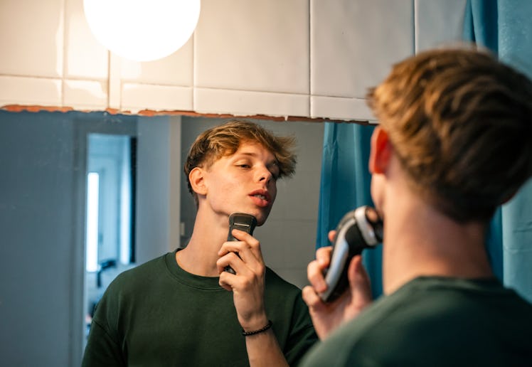 16-year-old young man shaves with a razor in front of the mirror in the bathroom. He is a handsome f...