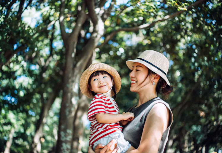 A loving young Asian mother carrying her cute little girl in arms, enjoying nature while spending qu...