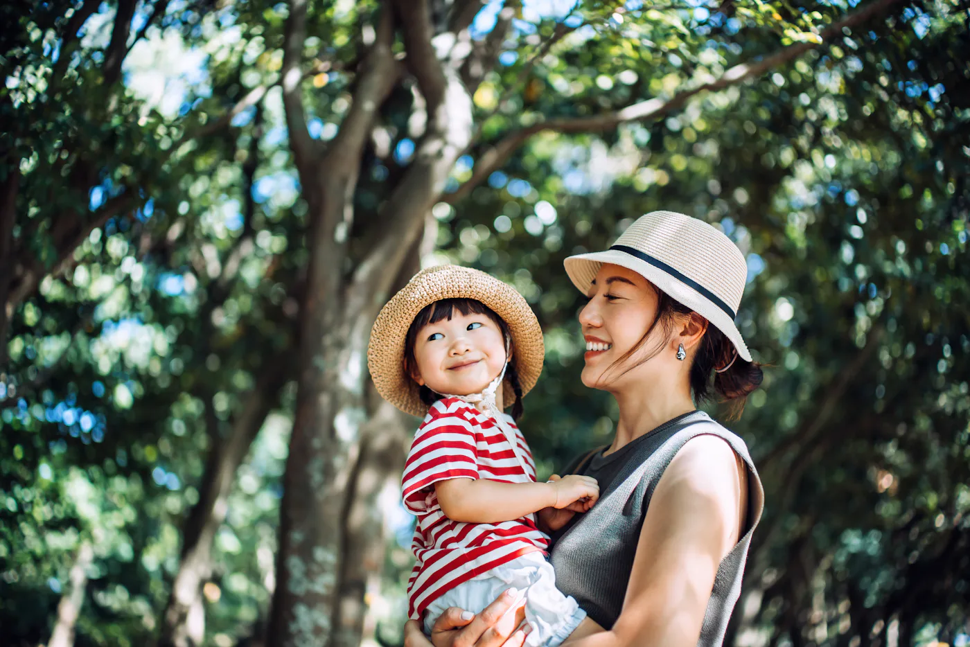 A loving young Asian mother carrying her cute little girl in arms, enjoying nature while spending quality mother-daughter moments together outdoors in park. Close life moments. Connecting to the nature. Sustainable lifestyle. Family love, care and bonding