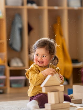 A joyful mixed-heritage toddler girl in a mustard sweater stacks wooden blocks in a warm daycare cla...