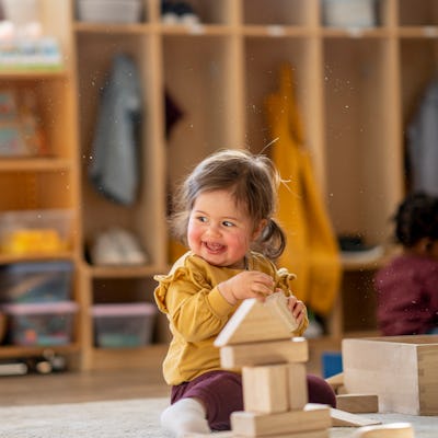 A joyful mixed-heritage toddler girl in a mustard sweater stacks wooden blocks in a warm daycare classroom. Colorful shelves and soft, natural light create a playful, learning-focused atmosphere.