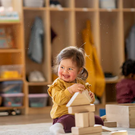 A joyful mixed-heritage toddler girl in a mustard sweater stacks wooden blocks in a warm daycare cla...