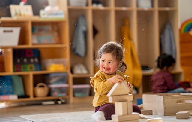 A joyful mixed-heritage toddler girl in a mustard sweater stacks wooden blocks in a warm daycare cla...