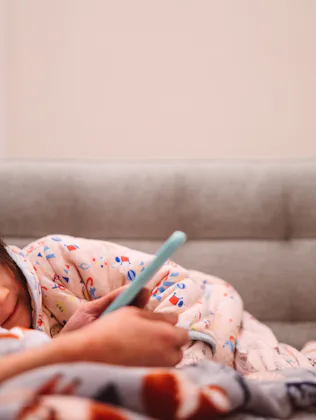 Lovely little girl using smartphone with her young mom while they are snuggling on the sofa at home.