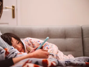 Lovely little girl using smartphone with her young mom while they are snuggling on the sofa at home.