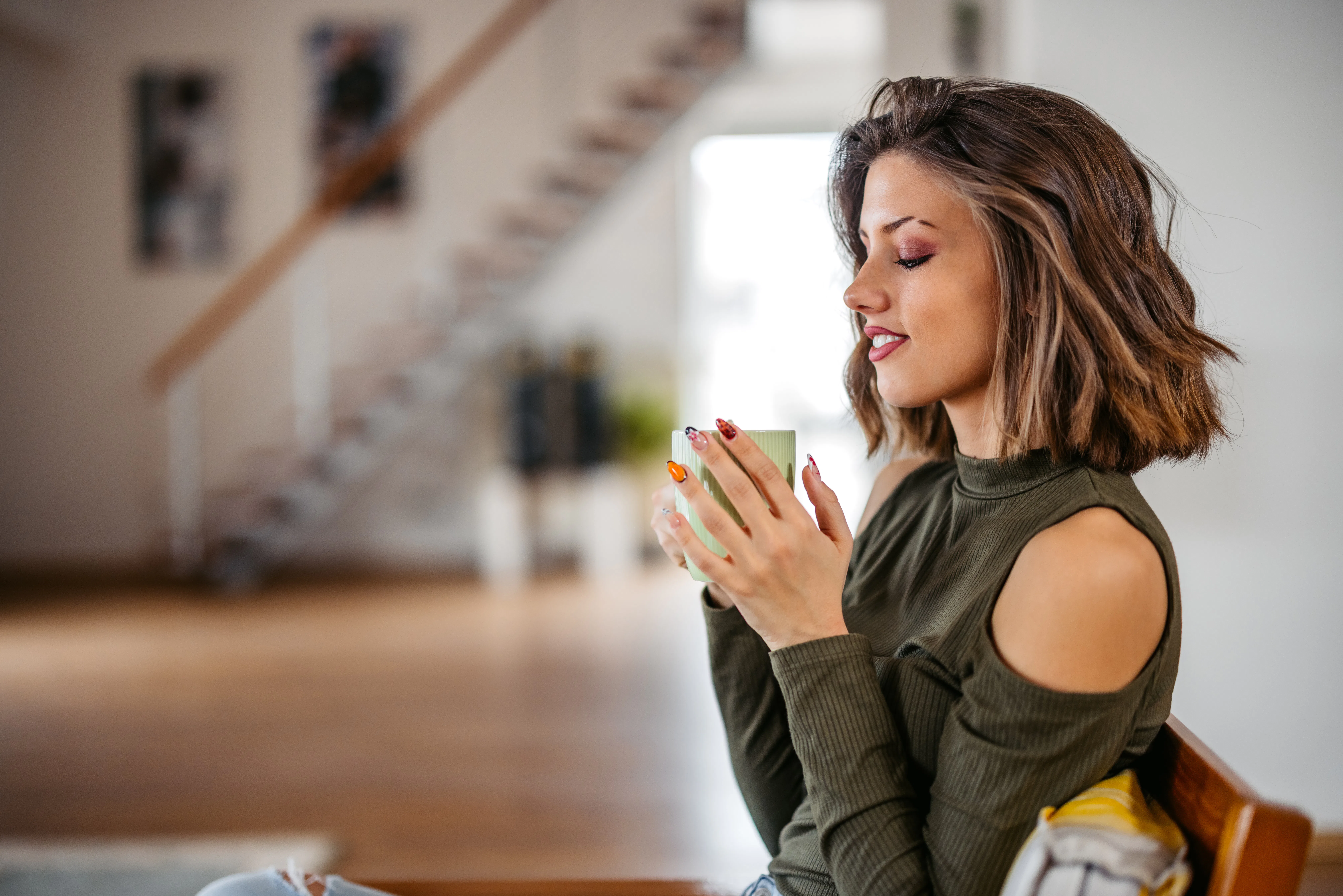Beautiful young woman sitting in the armchair in the living room at home and drinking coffee.