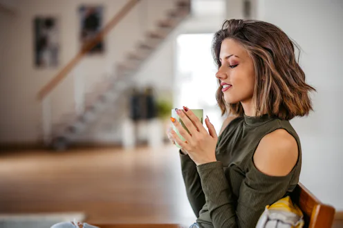 Beautiful young woman sitting in the armchair in the living room at home and drinking coffee.