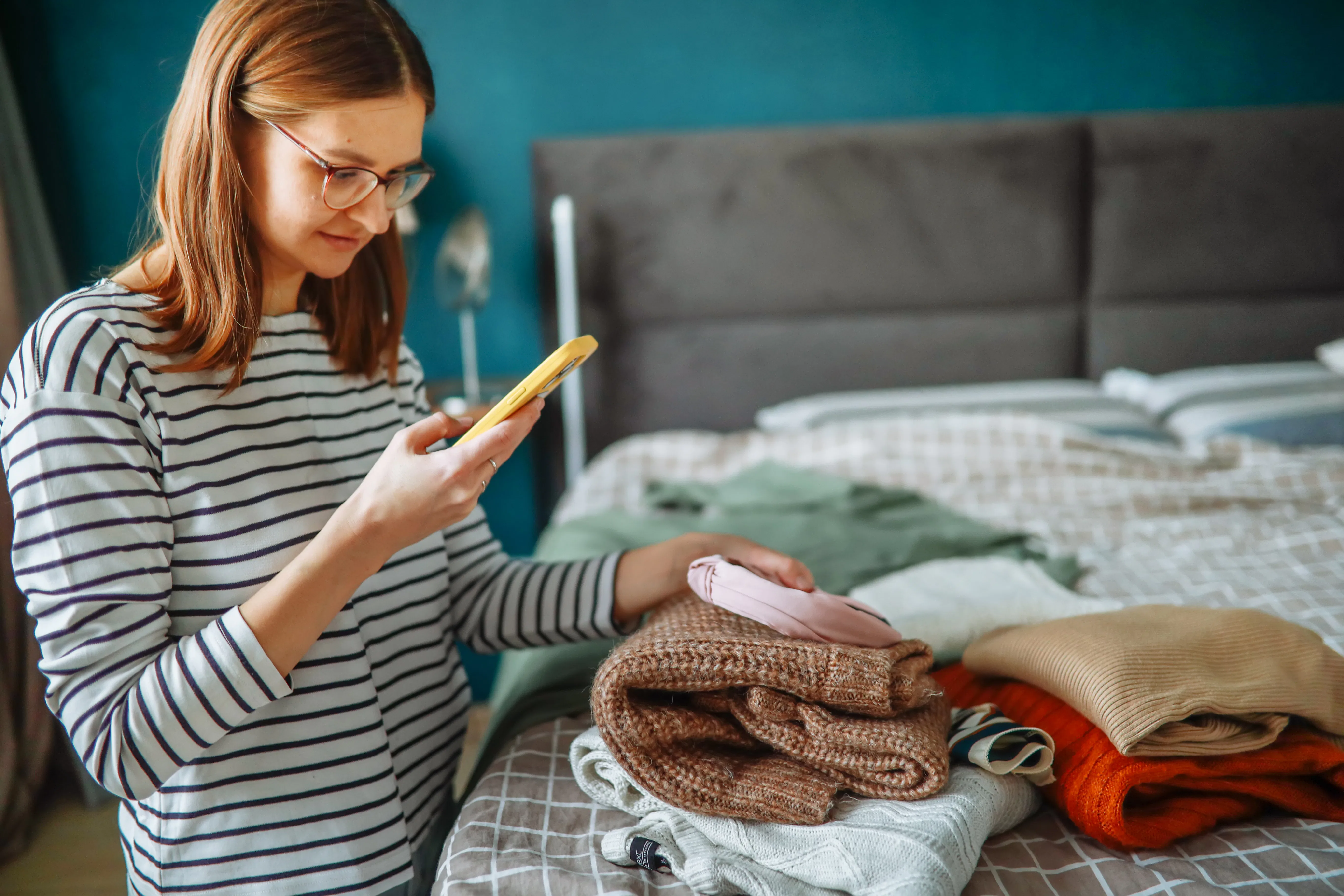 middle-aged woman in glasses photographs clothing laid out on a bed in a cozy bedroom using her smar...