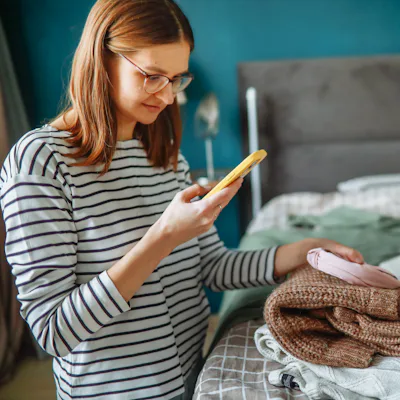 middle-aged woman in glasses photographs clothing laid out on a bed in a cozy bedroom using her smartphone, selling second-hand items and choosing clothes with the help of an online stylist at home.