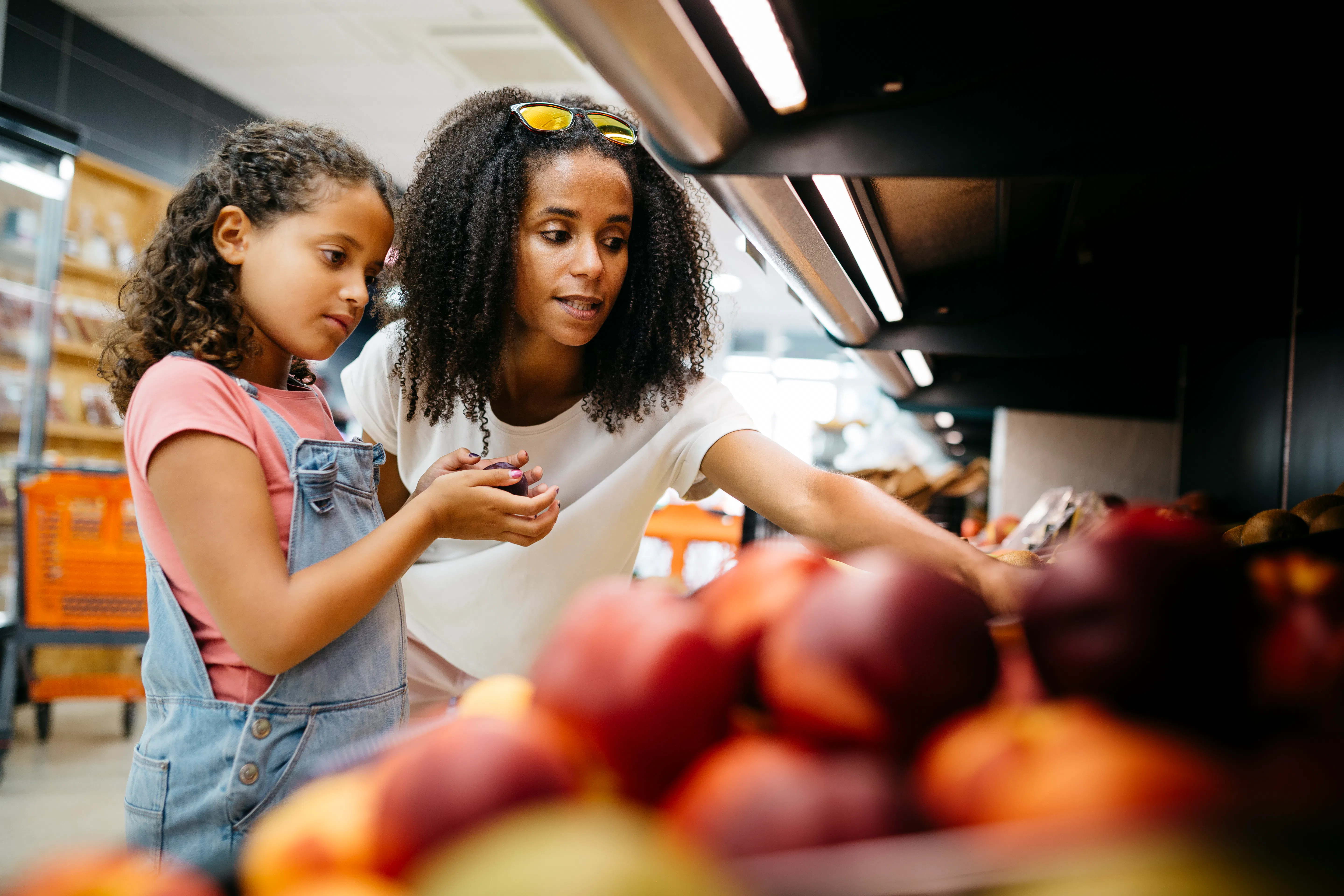 Black single mother and her daughter selecting fruits and vegetables from shelf while shopping in a ...