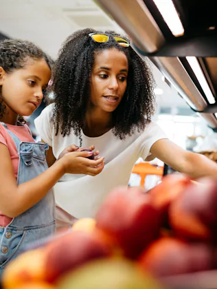 Black single mother and her daughter selecting fruits and vegetables from shelf while shopping in a ...