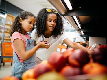 Black single mother and her daughter selecting fruits and vegetables from shelf while shopping in a ...