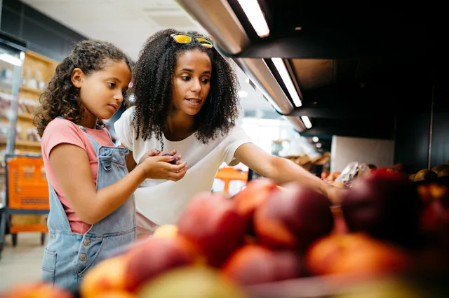 Black single mother and her daughter selecting fruits and vegetables from shelf while shopping in a ...