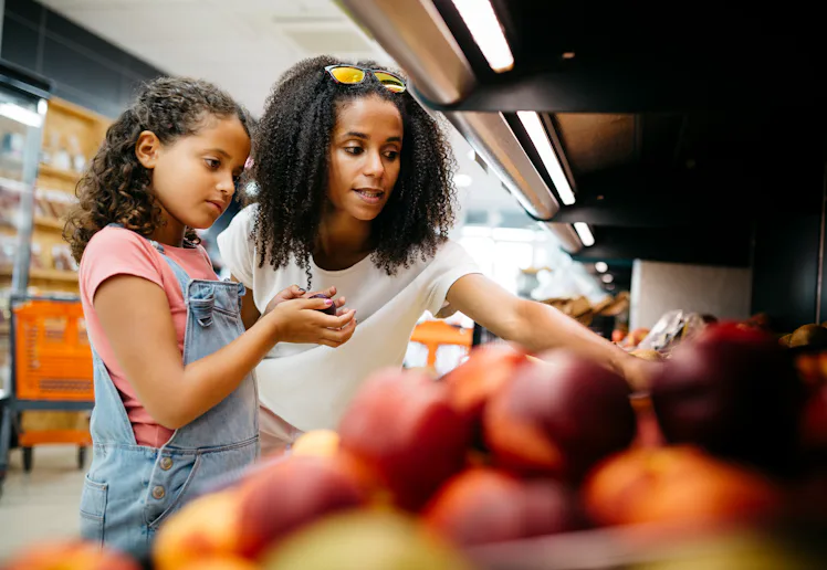 Black single mother and her daughter selecting fruits and vegetables from shelf while shopping in a ...