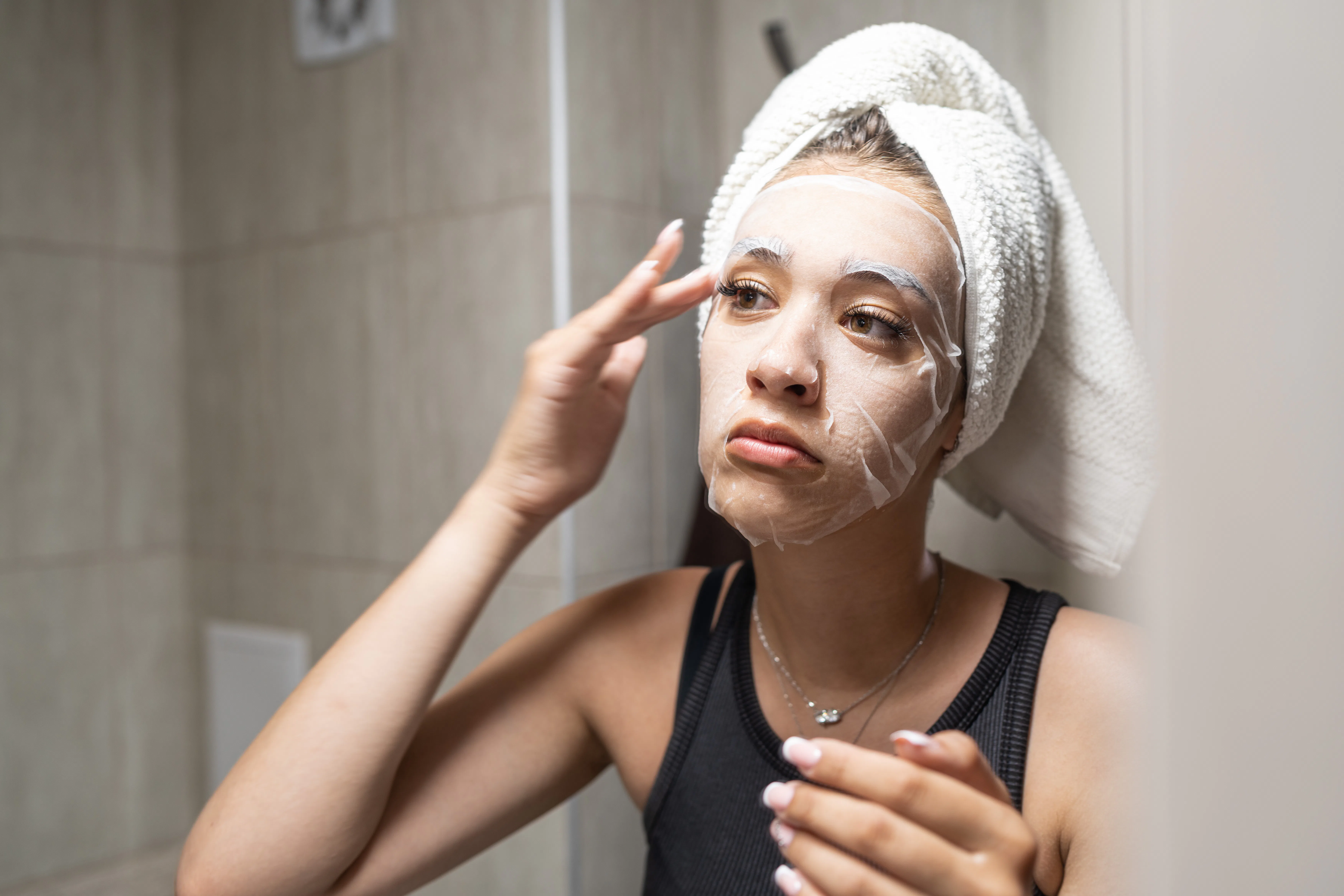 Young woman with a towel wrapped around her head carefully applying a hydrating sheet mask to her fa...
