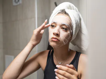 Young woman with a towel wrapped around her head carefully applying a hydrating sheet mask to her fa...