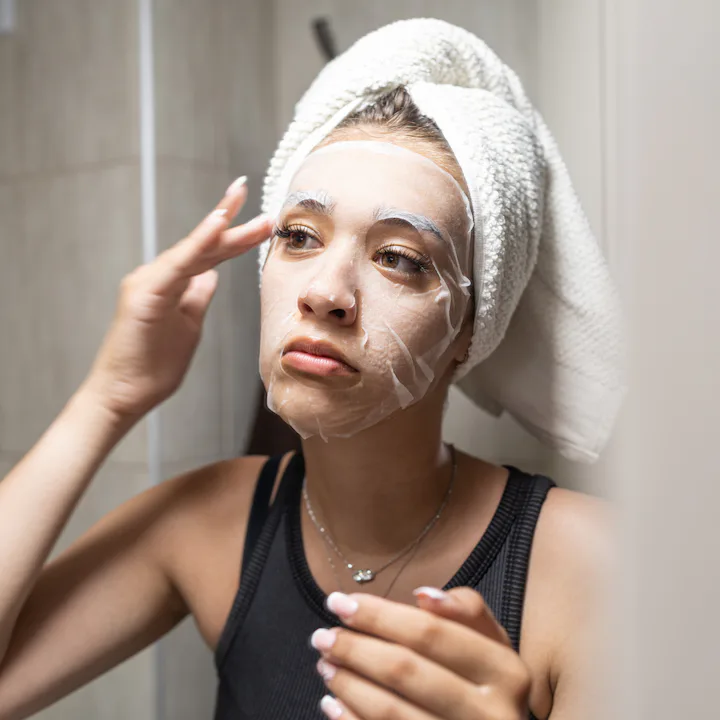 Young woman with a towel wrapped around her head carefully applying a hydrating sheet mask to her fa...
