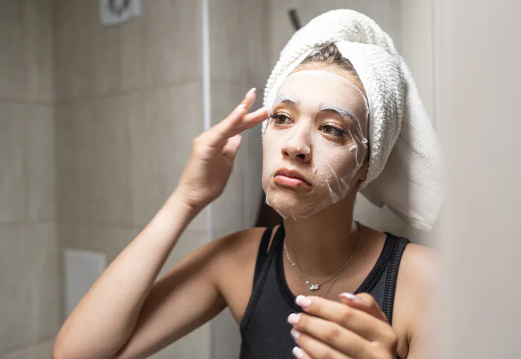Young woman with a towel wrapped around her head carefully applying a hydrating sheet mask to her fa...