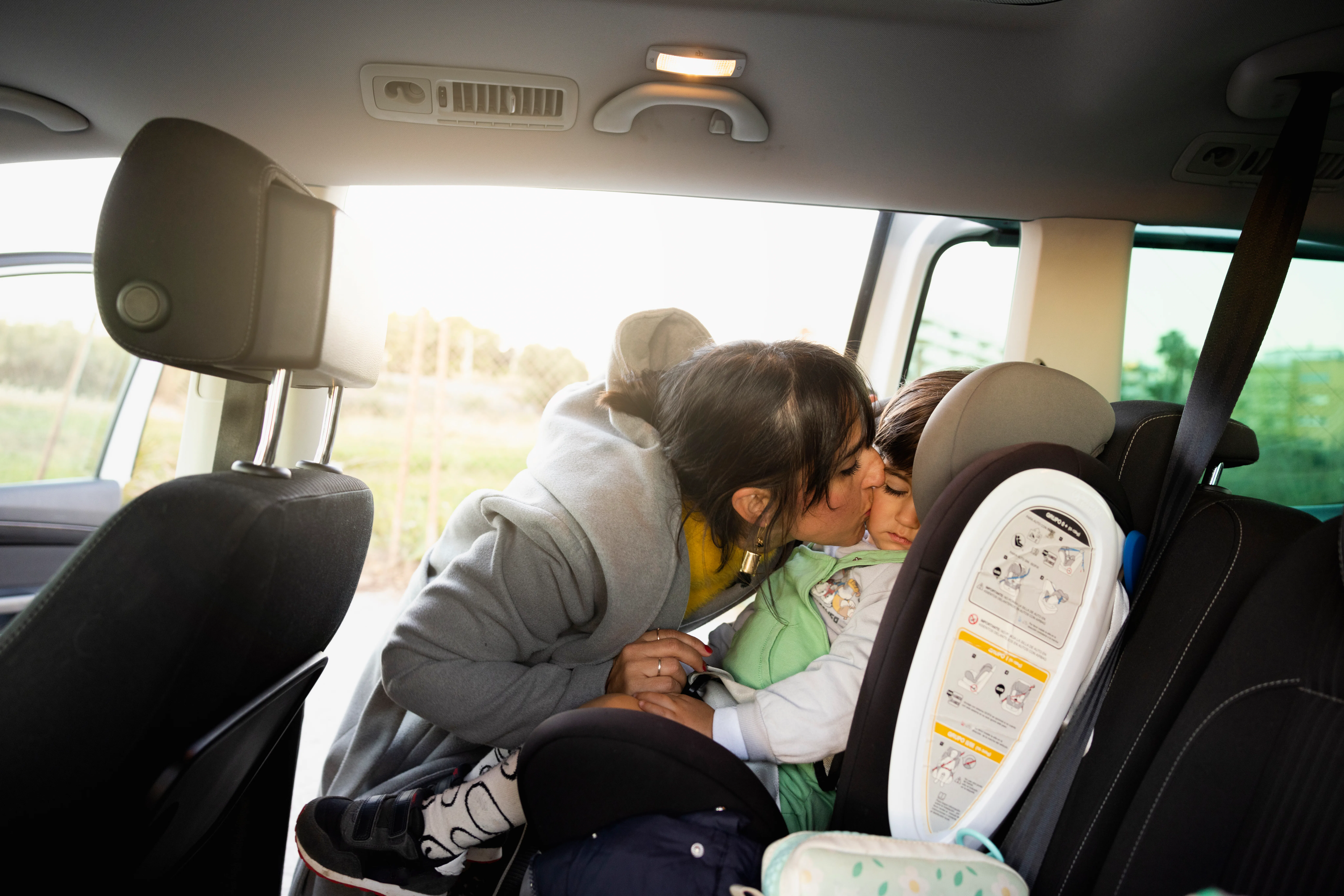 Mother kissing and securing her toddler boy buckled into his  baby safety car seat. Mother prepearin...