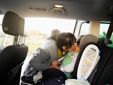 Mother kissing and securing her toddler boy buckled into his baby safety car seat. Mother prepearin...