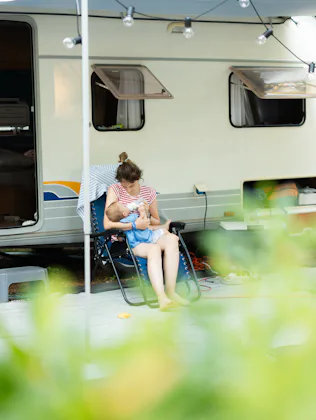 Mother bottle feeding her baby while sitting in front of a travel trailer at a campsite