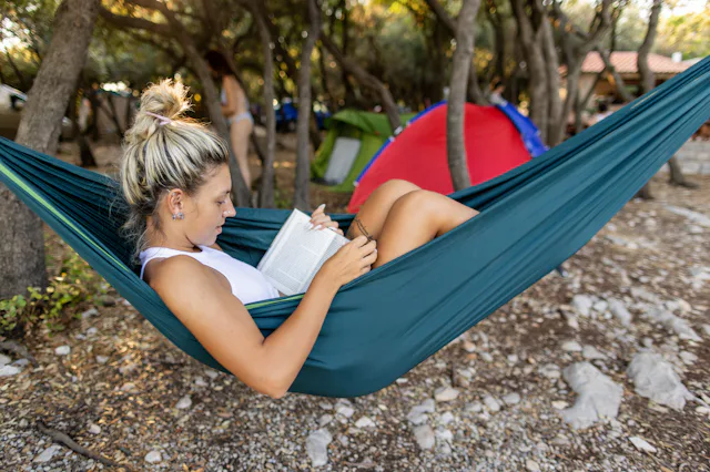 Young woman is camping in summer - she is lying in a hammock, reading a book and having a relaxing t...