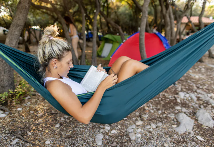 Young woman is camping in summer - she is lying in a hammock, reading a book and having a relaxing t...