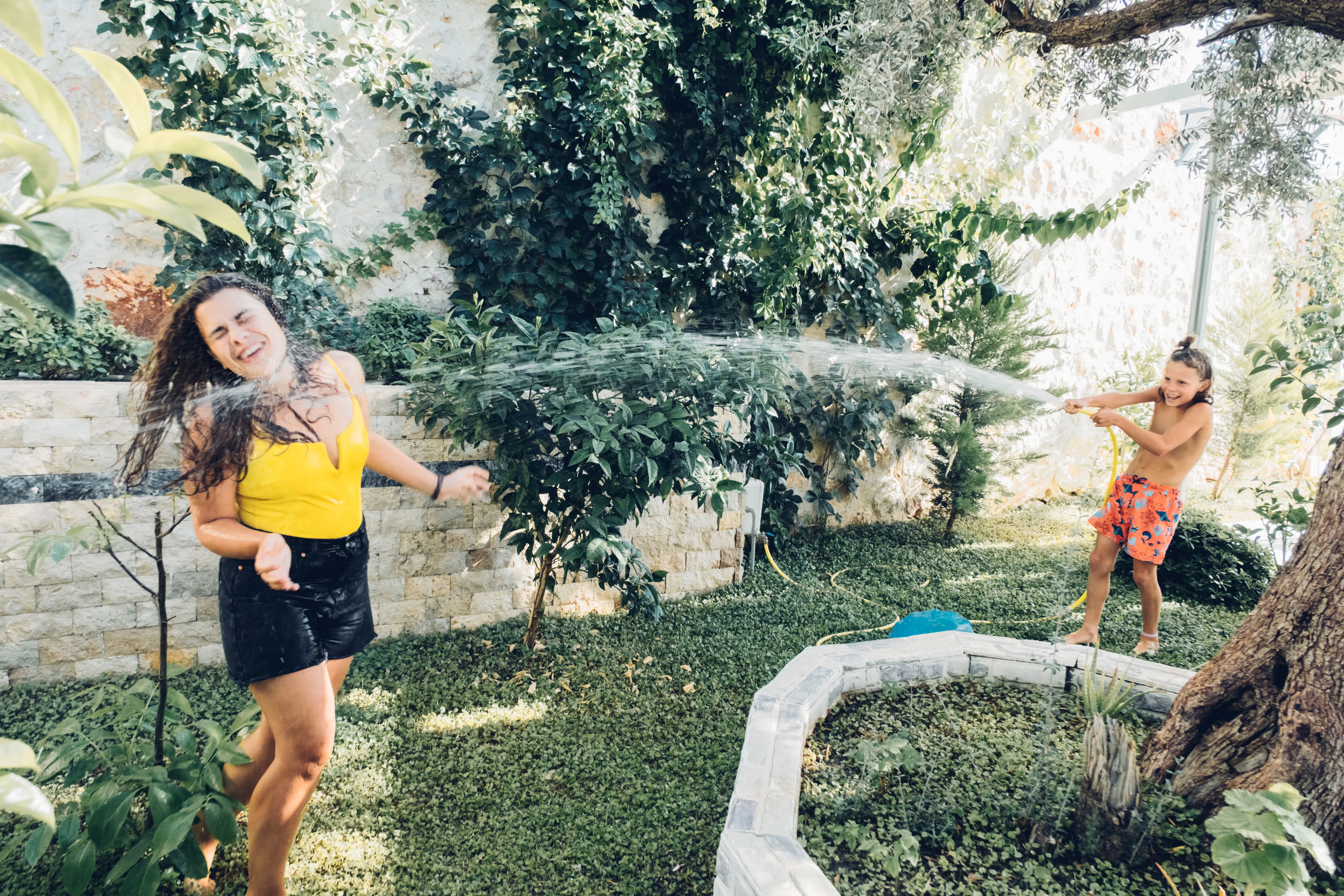 Happy young woman and cute child boy playing with water hose in garden. Weekend activity