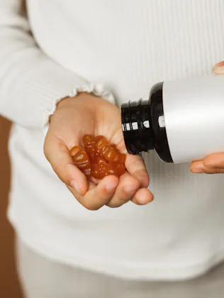 Small person in a light sweater pours brown gummy vitamins into their hand from a dark bottle, highl...