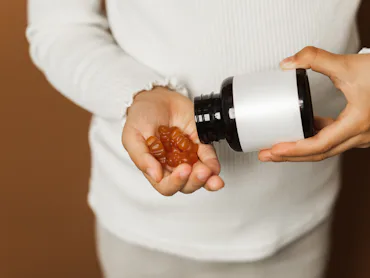 Small person in a light sweater pours brown gummy vitamins into their hand from a dark bottle, highl...