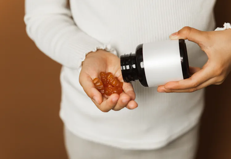Small person in a light sweater pours brown gummy vitamins into their hand from a dark bottle, highl...