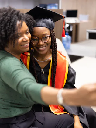 A proud mother takes a selfie with her daughter in graduation attire, capturing a joyful and memorab...