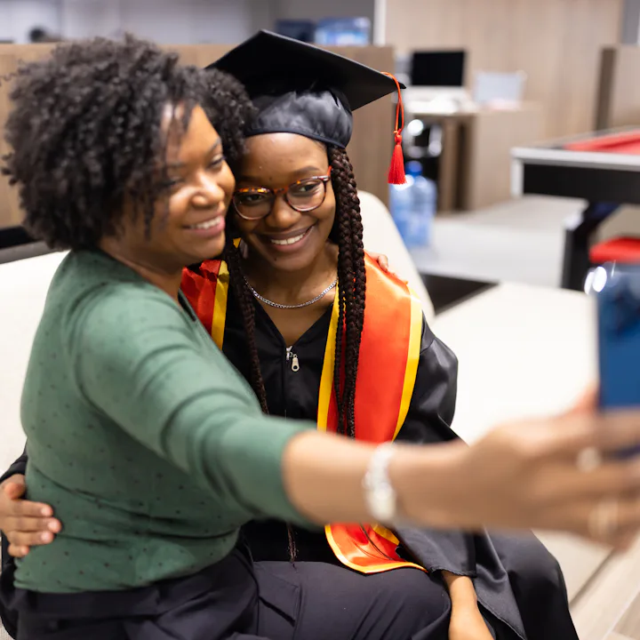 A proud mother takes a selfie with her daughter in graduation attire, capturing a joyful and memorab...